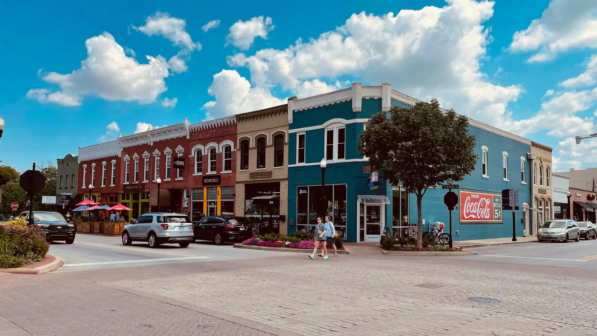 A wide angle shot of the Bentonville, AR downtown. A wide array of local small businesses are shown, with pedestrians walking to and from.