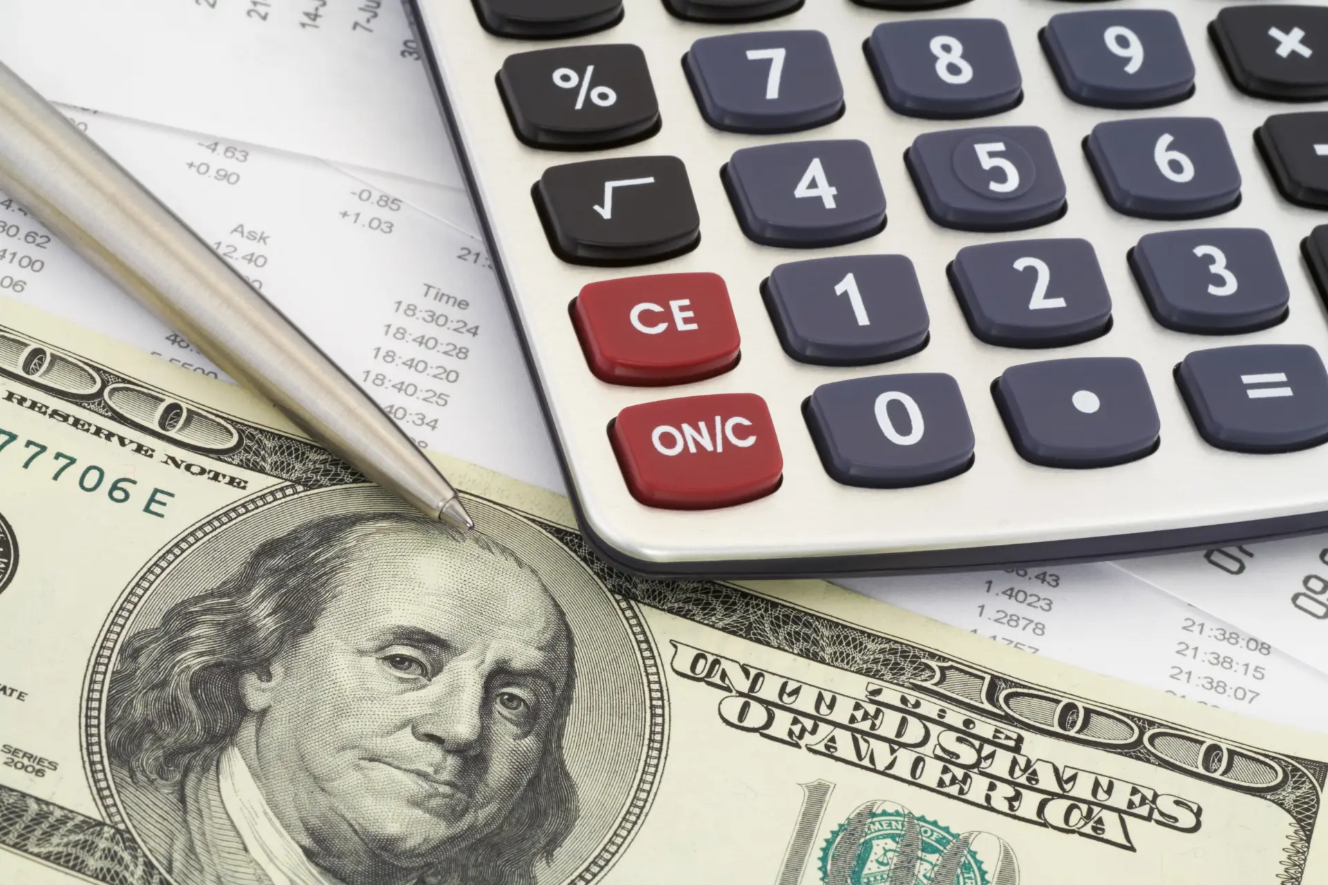 A close up photo of a calculator, pen, and hundred dollar bill laying on top of bills and receipts.